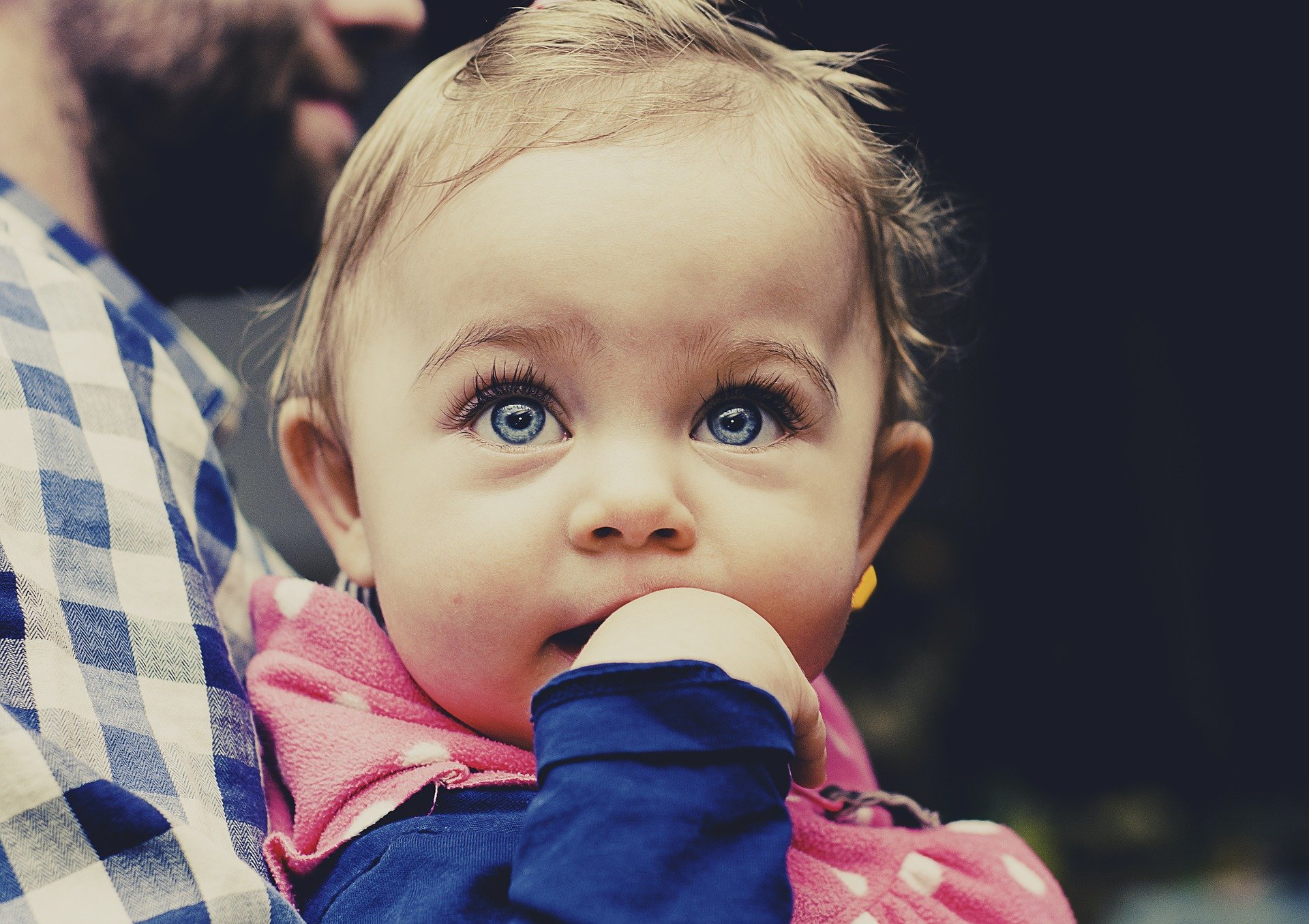 푸른 눈을 가진 외국 어린아이가 오른손을 입에 넣고 정면위를 바라보며 아빠품에 안겨있는 사진