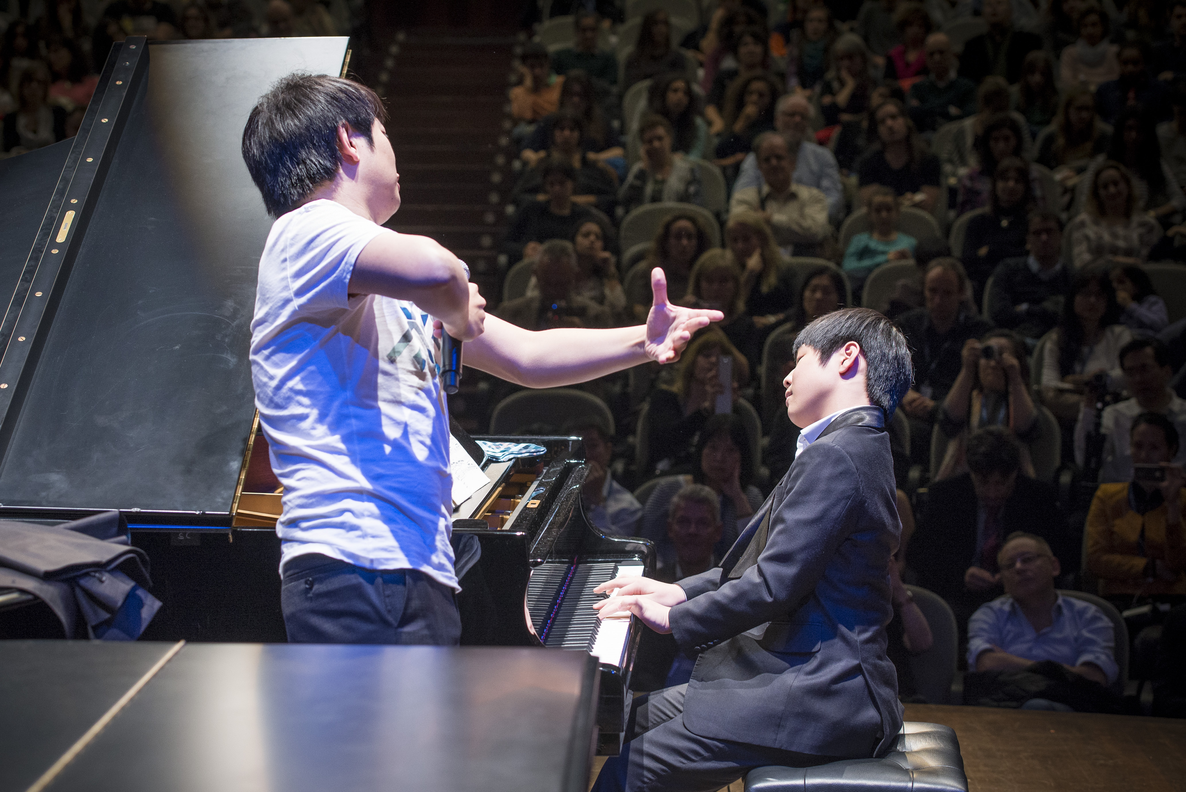 청중들 앞에서 피아노를 치는 소년과 마이크를 들고 솟짓을 하고 있는 청년의 사진