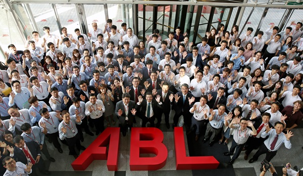  ABL글씨 조형물이 놓여있고 그 주위로 ABL 순레이 사장과 전 직원들이 모여 양손을 하늘로 흔들며 밝게 웃는 모습을 위에서 아래로 찍은 사진